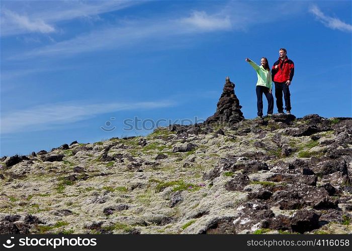 A man and a woman at the top of a mountain standing next to a cairn, the woman is pointiing to a distant horizon. Traditionally these rock piles were signposts but in recent times adding stones to them is deemed to bring good luck. Shot on location in Iceland.