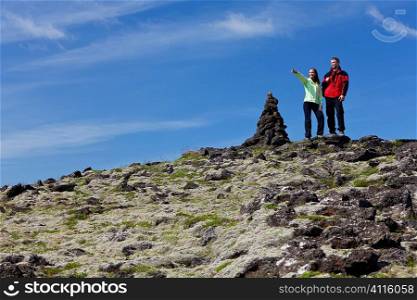 A man and a woman at the top of a mountain standing next to a cairn, the woman is pointiing to a distant horizon. Traditionally these rock piles were signposts but in recent times adding stones to them is deemed to bring good luck. Shot on location in Iceland.