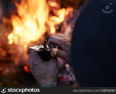 A man cuts a dried reindeer meat with a knife, camp fire on the background