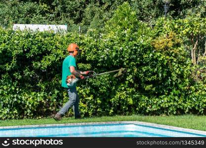 A man equipped with a brush cutter works on front yard with swimming pool. Garden maintenance concept