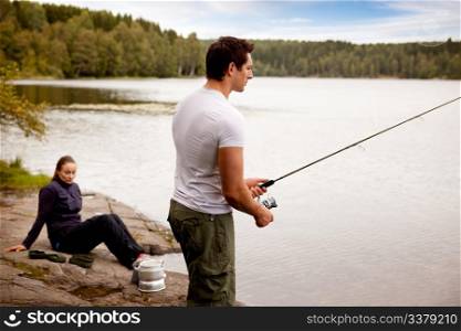 A man fishing on a lake with camping equipment and woman in background