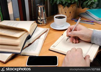 a man holds a pen and takes notes in a notebook on a desk in his office, surrounded by books, notebooks with a cup of black coffee