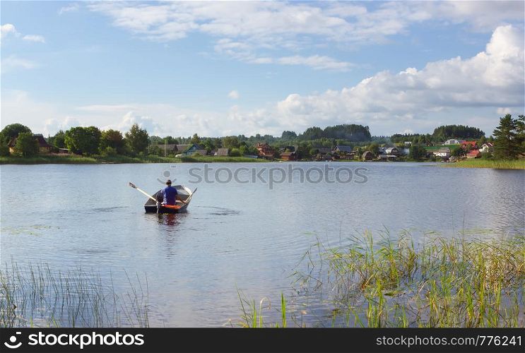 A man in a boat rowing to the village houses on a distant shore at sunny summer day. The concept of tourism, vacation, hobbies and outdoor activities - space for copy, selective focus. Lake Seliger, Russia.