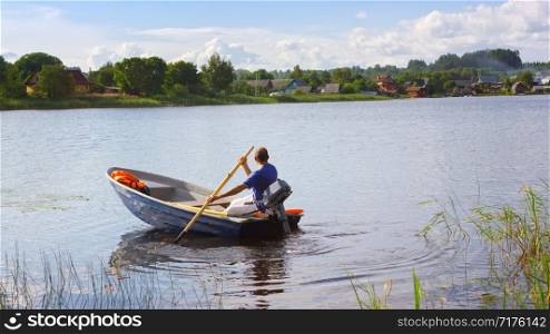A man in a motorboat with oars floating on the lake against the distant coast with village houses. The concept of tourism, vacation, hobbies and outdoor activities. Space for copy, selective focus. Summer day at Seliger Lake, Russia.