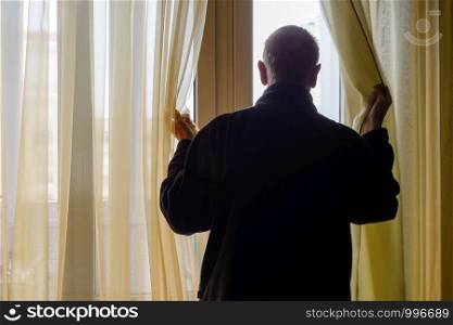 A man in black moves the curtain aside to look out trough the window