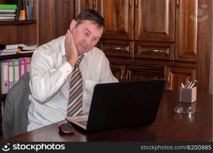 A man in his midlife sits in his stydy in front of a laptop while rubbing his neck as he stresfully looks at the screen.