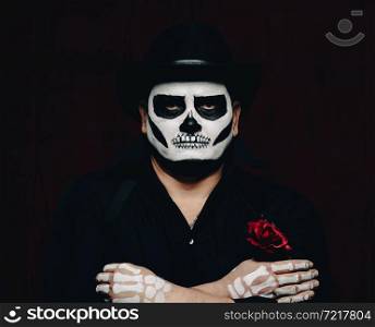 a man is painted with a skeleton in a black hat and shirt, arms crossed on his chest. Black background