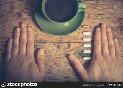 A man is resting his hands on a table with a cup of coffee and some pills