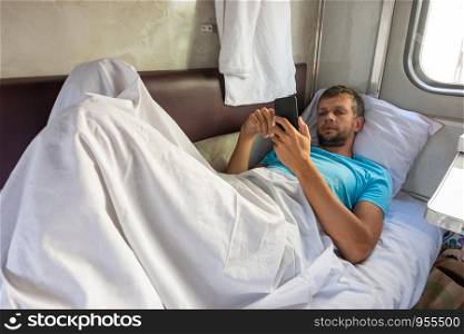 A man lies on the bottom shelf in a train car with a phone in his hands