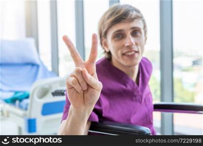 a man patient sit in a wheelchair Abstract blur with focus show showing lifts two fingers up fighting with illness up with smiley face very good symptom in hospital background.