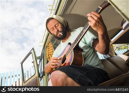 A man plays the guitar while sitting in his classic combi van and smoking a cigarette.