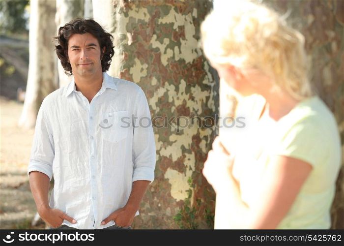 a man posing and a blonde woman hidden behind a tree watching him in secret