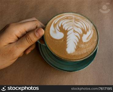 A man&rsquo;s hand holds a cup of cappuccino which sits on craft paper, a first-person view. A man&rsquo;s hand holds a cup of cappuccino, a first-person view