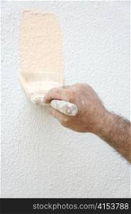 A man&rsquo;s paint-spattered hand holding a paintbrush and painting a house.