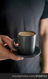 A man’s hand holds a black cup of hot espresso coffee against the background of a black T-shirt.. A man’s hand holds a cup of hot espresso coffee against the background of a black T-shirt.