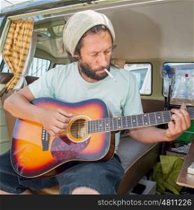 A man sits on the back seat of his camper van and plays the guitar while smoking a sigarette.