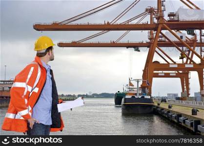A man wearing a safety coat and a hard hat inspecting the huge cranes at a container harbor