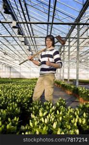 A man with a broom in his hand giving a thumbs-up inside a glasshouse, surrounded by potted plants
