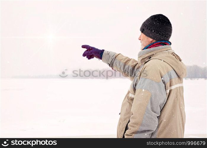 A man with a wool cap, a waterproof jacket and a wool cap is indicating something with the finger close to the Dnieper river during a cold and sad gray winter morning under the snow