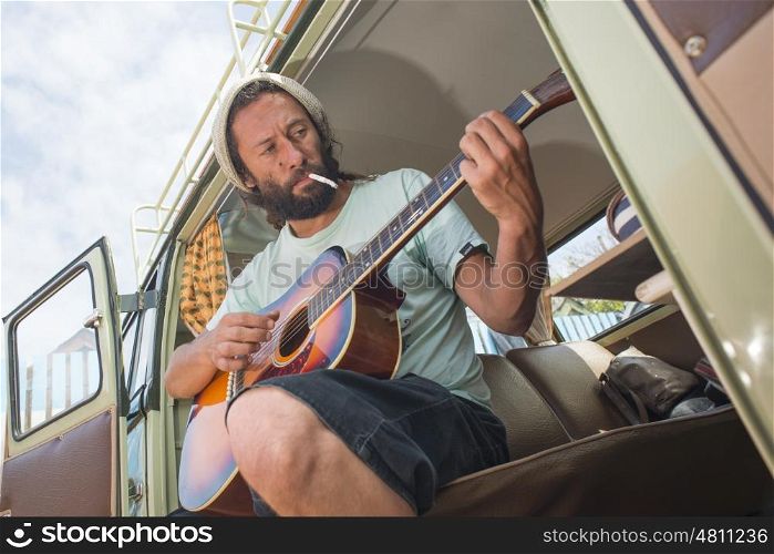 A mans sits with a cigarette in his mouth at the side door of his classic combi van while playing the guitar.