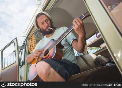 A mans sits with a cigarette in his mouth at the side door of his classic combi van while playing the guitar.