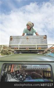 A mans stands on the roof of his camper van as he packs a big metal crate on the roof rack of his camper van as viewed from the back of the vehicle.