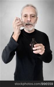 A mature man is drinking a spoon of cough syrup to cure his sore throat and bronchitis