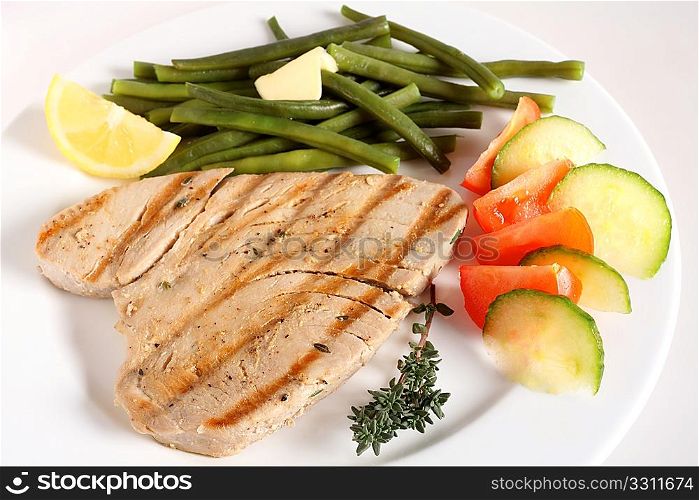 A meal of grilled tuna steak with a salad of tomato and cucumber and green beans, garnished with a sprig of thyme and a lemon wedge