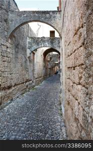 A medieval cobblestone alley in Rhodes Old Town (UNESCO World Heritage Site), Greece, with arches and stone facades.