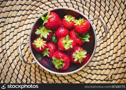 A metallic bowl of red ripe strawberries on a circular straw mat, viewed from above / flat lay