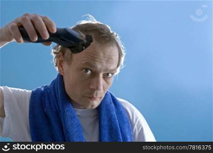 A middle aged man dressed in a white t-shirt, shaving his own hair with a clipper