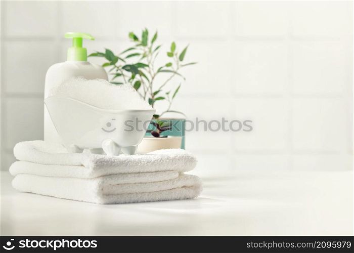 A miniature bubble bath, shampoo, flowers and white towels on bathroom countertop, copy space