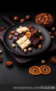 A mix of several types of delicious sweet chocolate broken into cubes on a black plate on a dark concrete background