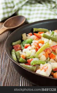 A mixture of assorted frozen vegetables in black pan ready for cooking on brown wooden table with wooden spoon. Mixture of frozen vegetables on brown wooden background