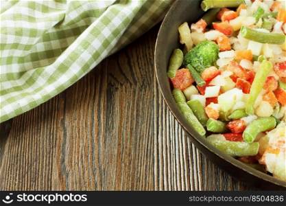 A mixture of assorted frozen vegetables in black pan ready for cooking on brown wooden table with copy space for text. Mixture of frozen vegetables on brown wooden background