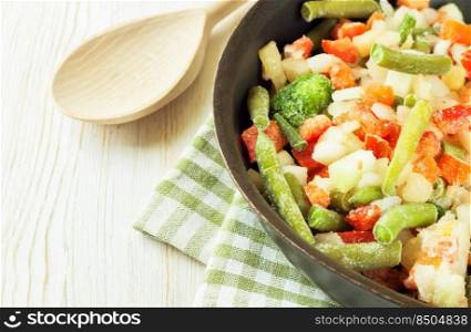 A mixture of assorted frozen vegetables in black pan ready for cooking on white wooden table with copy space for text. Mixture of frozen vegetables on white wooden background