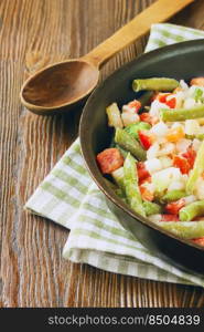 A mixture of assorted frozen vegetables in black pan ready for cooking on brown wooden table with wooden spoon. Mixture of frozen vegetables on brown wooden background