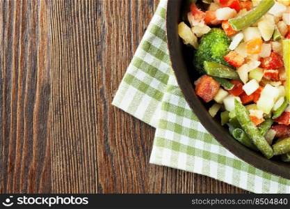 A mixture of assorted frozen vegetables in pan ready for cooking on brown wooden table. Top view, flat lay with copy space for text. Mixture of frozen vegetables on brown wooden background