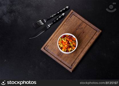 A mixture of chopped vegetables paprika, corn, peas in white plate against a dark concrete background. A mixture of chopped vegetables paprika, corn, peas in white plate