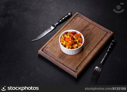 A mixture of chopped vegetables paprika, corn, peas in white plate against a dark concrete background. A mixture of chopped vegetables paprika, corn, peas in white plate