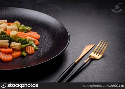 A mixture of vegetables  carrots, small heads of corn, asparagus beans steamed on a black ceramic plate