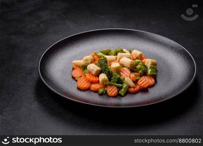 A mixture of vegetables: carrots, small heads of corn, asparagus beans steamed on a black ceramic plate