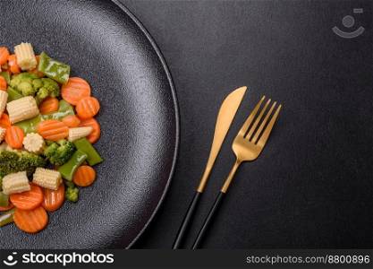 A mixture of vegetables: carrots, small heads of corn, asparagus beans steamed on a black ceramic plate