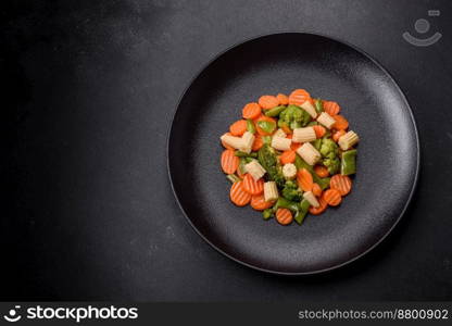 A mixture of vegetables  carrots, small heads of corn, asparagus beans steamed on a black ceramic plate