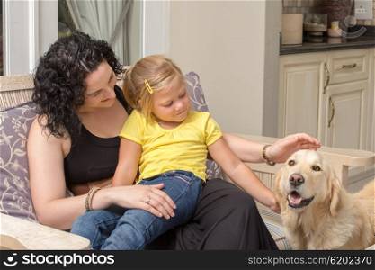 A mother and her daughter pat the golden retriever dog while sitting on a bench on their patio.
