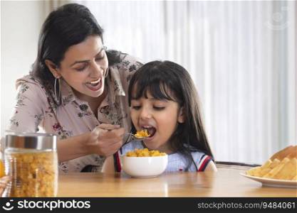 A MOTHER HAPPILY GIVING A SPOONFUL OF CORNFLAKES TO DAUGHTER