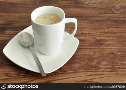 A mug of vegetable soup displayed with a spoon and plate on a wooden background