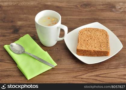 A mug of vegetable soup displayed with bread, a green napkin and a spoon on a wooden background