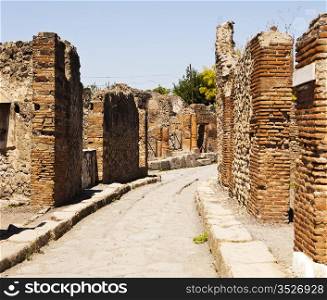 A narrow street in Pompeii winds between old brick walls of ancient homes in a residential area.