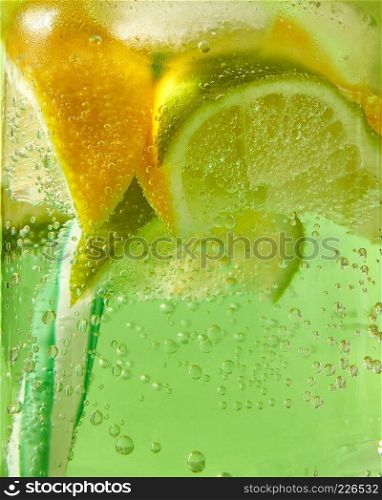 A natural fruit background with macro lime slices, lemon and a colored drinking plastic straw in a glass jar with aerated gassed bubbles. Concept of cold alcoholic or non-alcoholic summer drinks.. Close-up of sparkling fresh lemonade with ice, slices of lime, lemon and colored plastic straws in glass with large bubbles of air.
